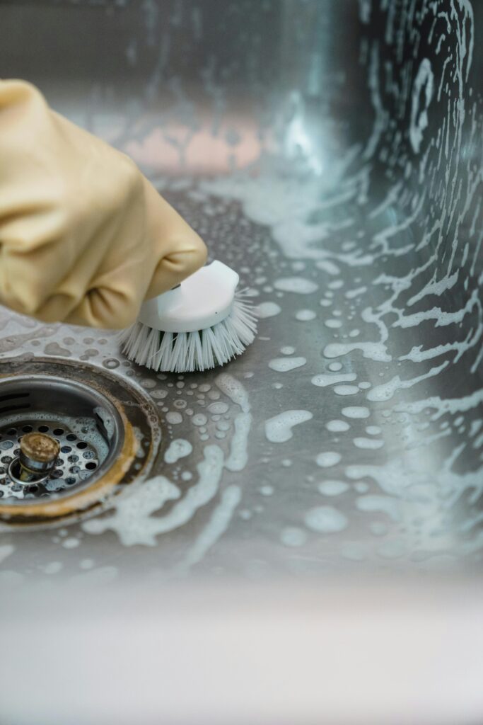 Close-up of a hand in gloves scrubbing a soapy kitchen sink with a brush.